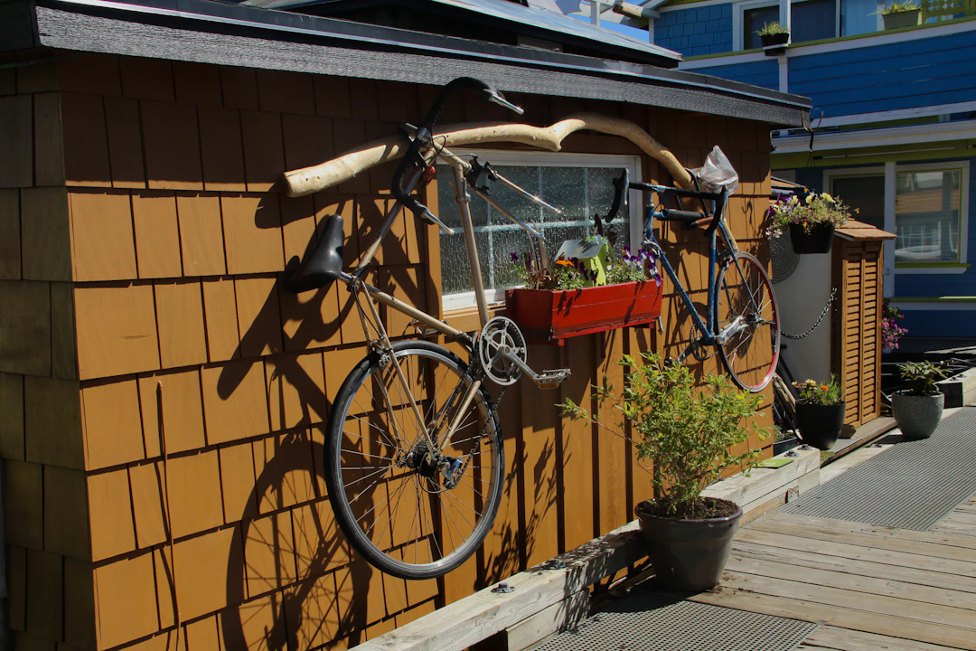Two bicycles mounted vertically on the exterior wall of a small wooden building. A rectangular red planter box with purple and yellow flowers sits between the bicycles below a window. Additional potted plants decorate the walkway in front, and a blue building is visible in the background.