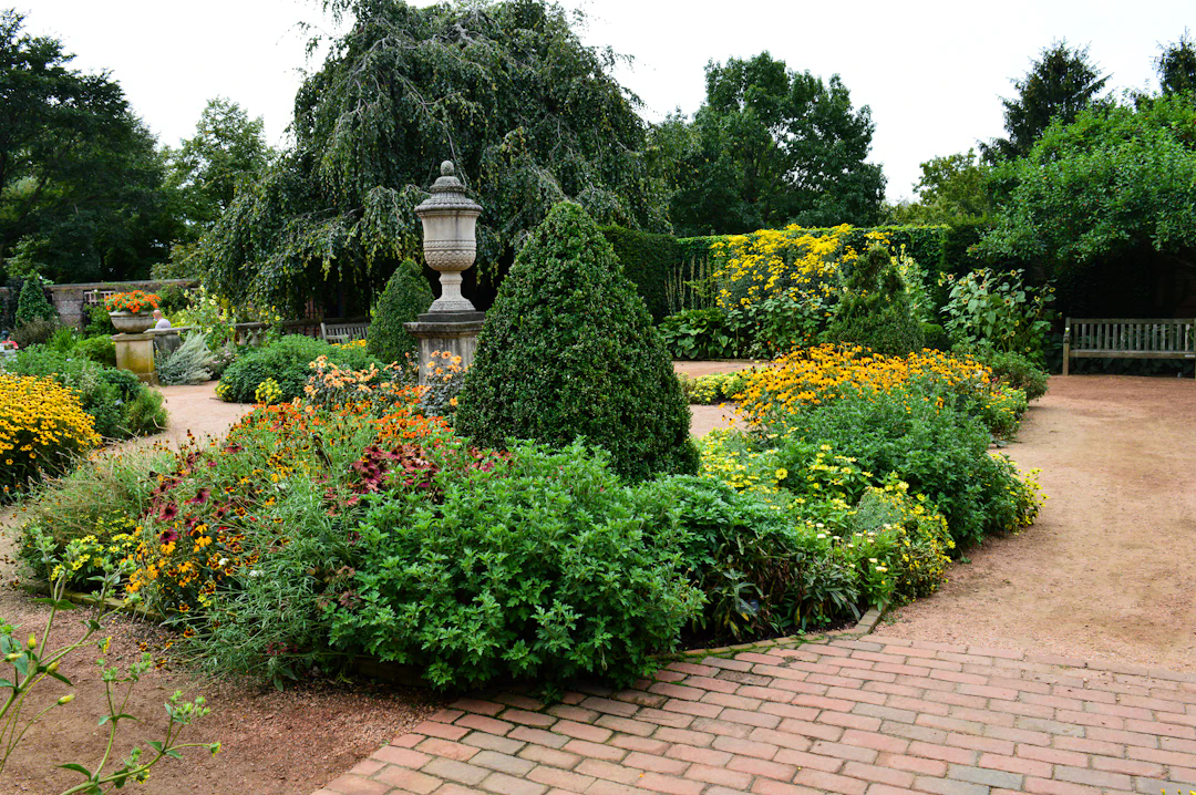 A beautifully landscaped formal garden featuring a variety of flowering plants and shrubs arranged in symmetrical beds. There are neatly trimmed bushes, vibrant yellow and orange flowers, and a stone urn at the center of a paved pathway. Benches and larger leafy trees border the garden, creating a peaceful and inviting atmosphere.