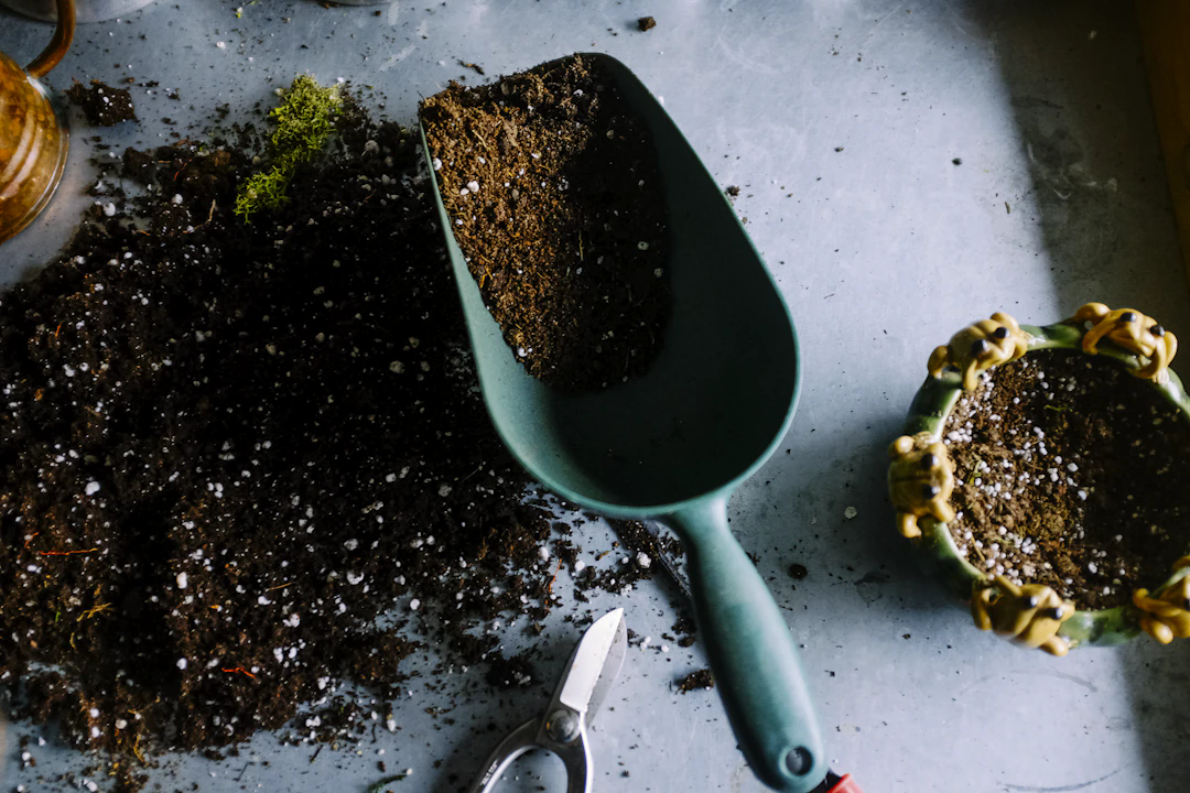 A close-up of a gardening scene showing a green hand trowel filled with soil, scattered soil on a surface, a pair of pruning shears, and a small decorative plant pot with frog designs on the rim, partially filled with soil.