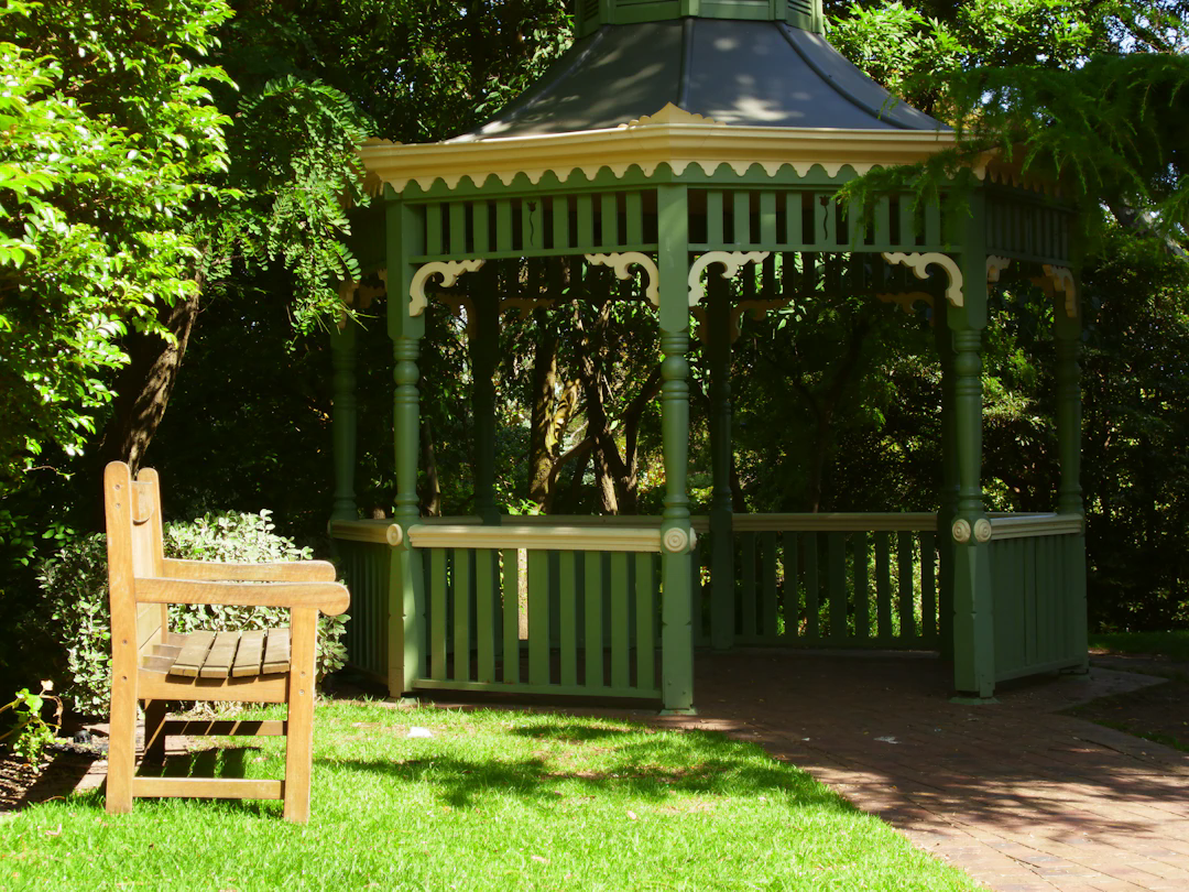 A green wooden gazebo with decorative trim is situated in a park or garden. In the foreground, there is a wooden bench placed on the grass. The gazebo is surrounded by lush green trees and plants, and sunlight creates dappled shadows on the ground.