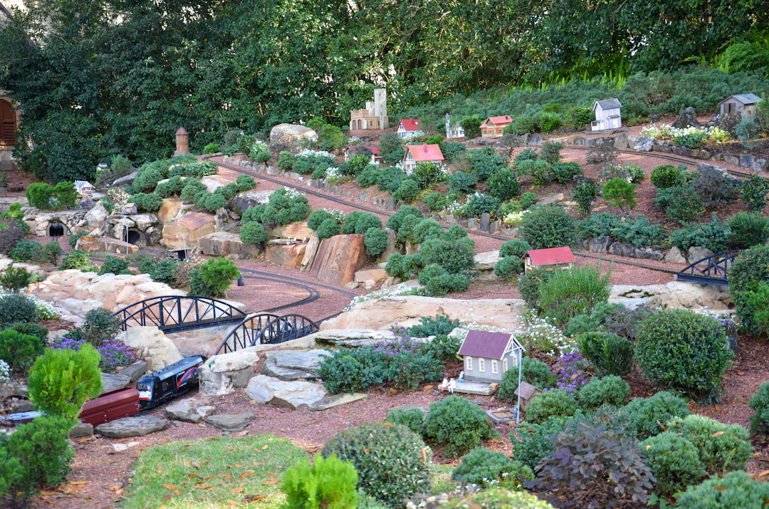 A detailed miniature garden railway landscape featuring a model train on tracks, small bridges, tunnel entrances, model houses, and abundant greenery resembling trees and bushes.