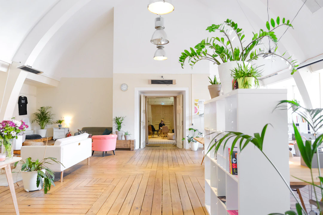 A bright, modern office space with high arched ceilings and wooden floors. The area is decorated with many green potted plants, white furniture including a sofa and bookshelves, and a pink armchair. A few people are seen working in a meeting room at the back. Natural light fills the space through large windows on the right.