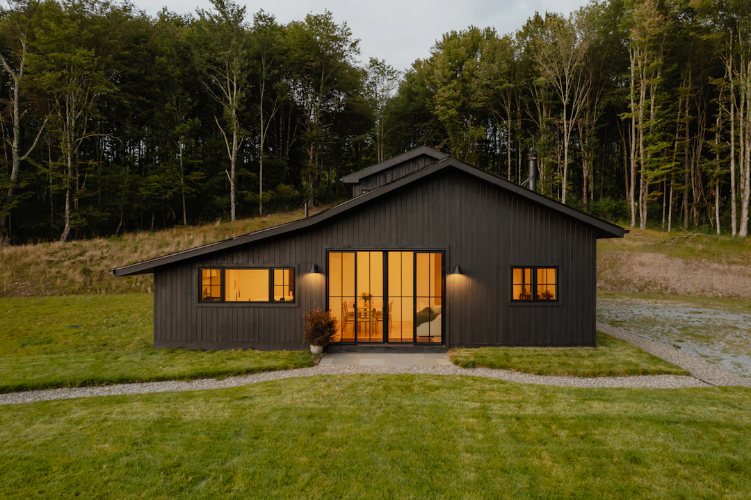 A modern, single-story dark wood house with large windows and warm interior lighting, situated on a grassy plot with a gravel pathway and surrounded by dense forest.
