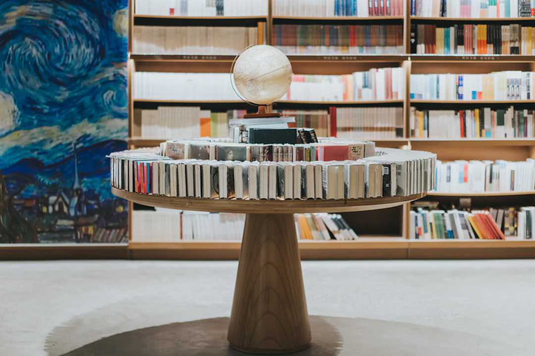 A modern library or bookstore interior featuring a round wooden table with books arranged standing up around the edge in a circle. On top of the table, there are stacked books and a globe. In the background, there are shelves filled with more books and a large mural resembling Van Gogh's Starry Night.