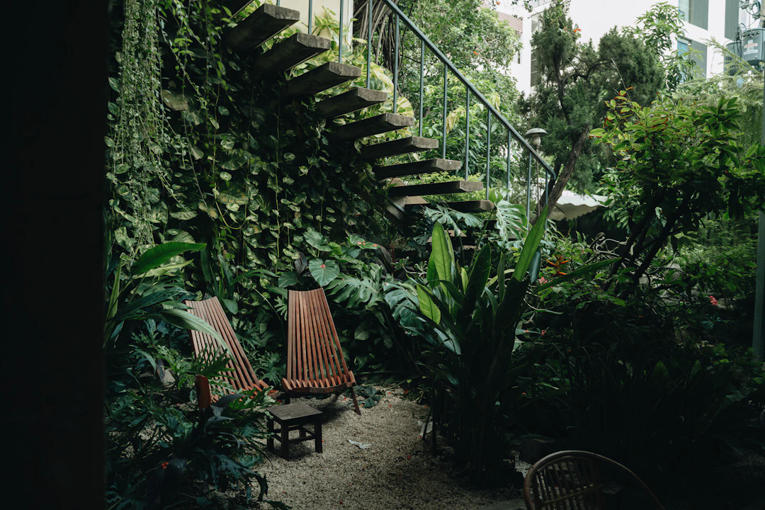A lush, green outdoor space featuring two wooden lounge chairs and a small wooden table, surrounded by various tropical plants. There is an open wooden staircase with a metal railing leading upwards, and the area looks secluded and serene, ideal for relaxation.