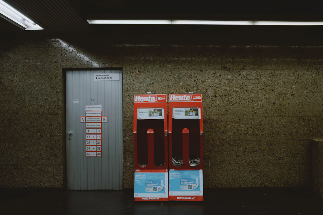 A dimly lit interior of a subway station or an underground area with a textured brown wall. On the left, there's a gray metallic door with signage and a list of numbers and texts in German. To the right of the door are two red and white newspaper stands labeled 'Heute' with slots for free newspapers, some of which are still inside. Fluorescent lights are visible on the ceiling.