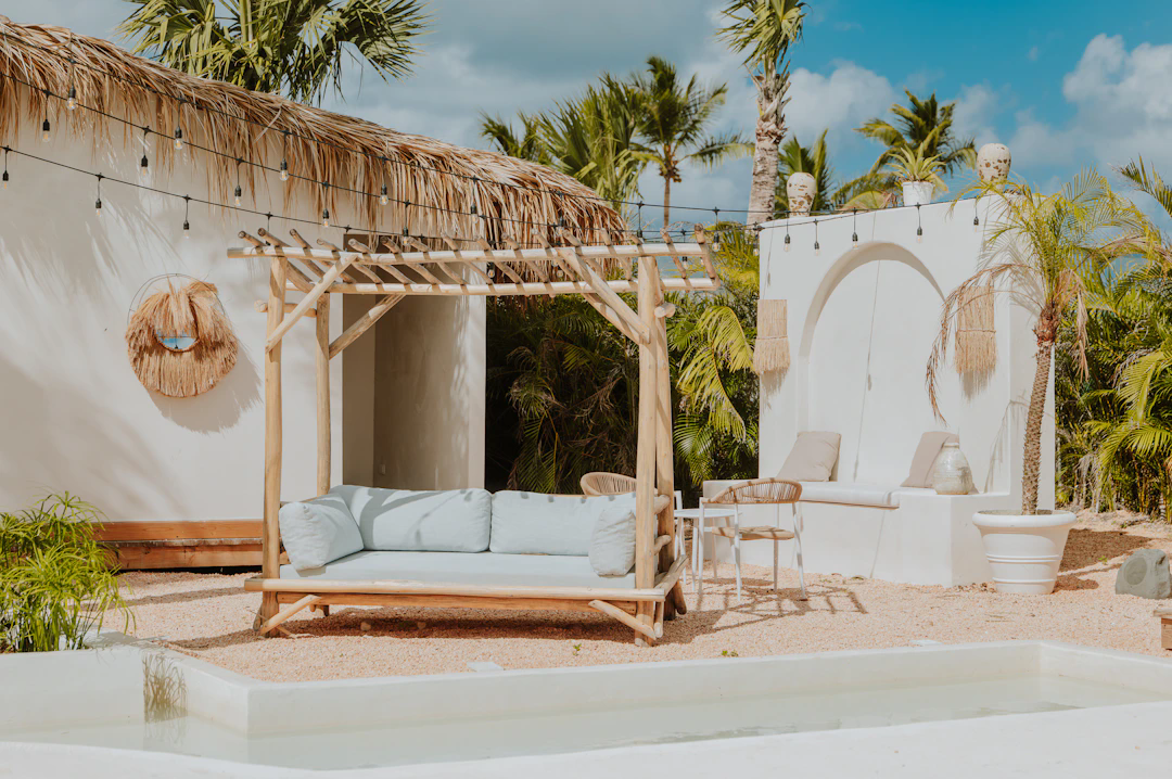 A stylish outdoor lounging area featuring a light wooden pergola with a cushioned sofa underneath, tropical palm trees in the background, a thatched roof, and decorative elements such as hanging lights and straw wall art. There are also white walls, patio furniture, and a shallow water feature in the foreground, all set under a partly cloudy sky.