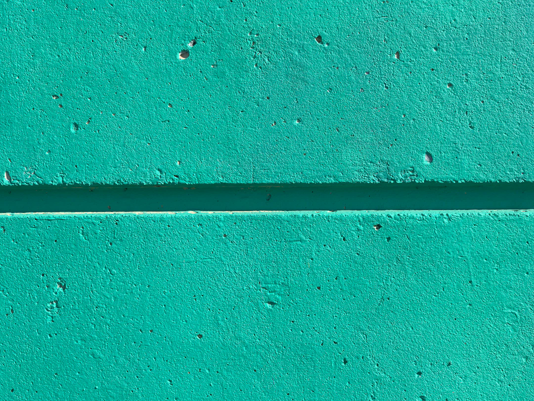 A close-up view of a turquoise-colored concrete wall with a horizontal groove running through the middle and small holes and texture variations visible.