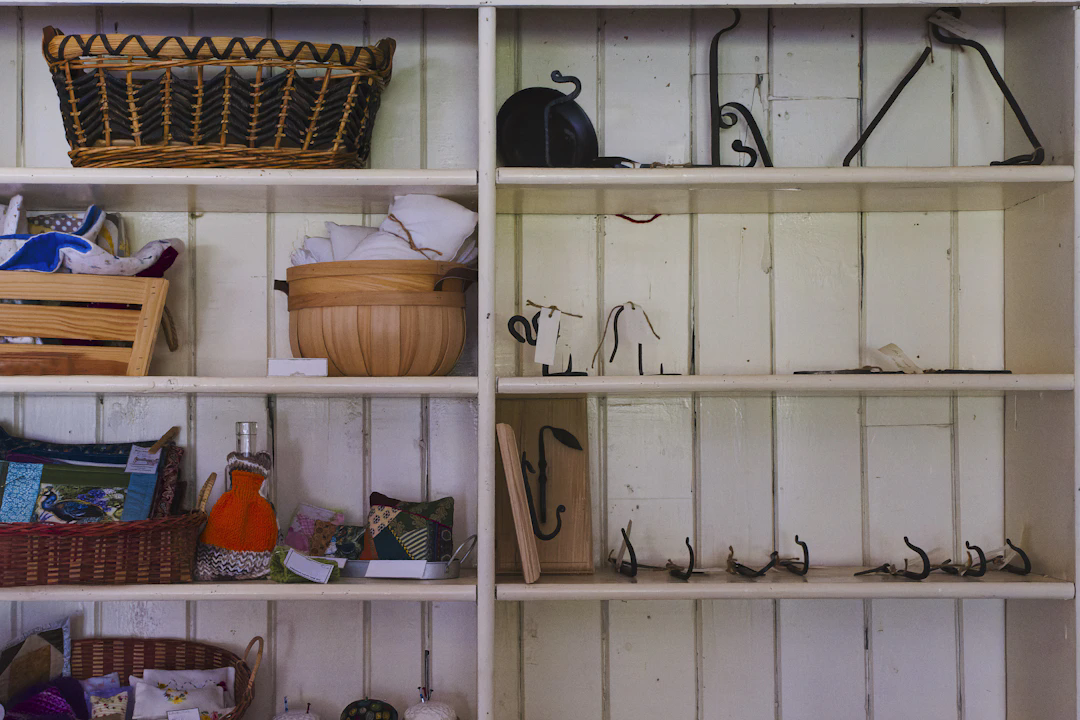 A set of wooden shelves attached to a white-paneled wall. The left shelves contain baskets, fabric, and handmade crafts including pillows, knitted items, and cloth pieces. The right shelves display metal hooks, a wooden bowl, and a black kettle, with many shelves partially empty except for iron hooks and hardware.