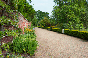 A well-maintained garden pathway bordered by lush green plants, blooming flowers, and neatly trimmed hedges. On the left, there is a red brick wall with flowering plants growing against it. On the right, ornamental trees and topiary bushes decorate the formal garden. The walkway is made of fine gravel, and a park bench is visible further down the path.