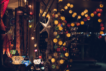 A cozy indoor market or shop decorated with colorful string lights and ornate lanterns. Yellow, red, green, and white round lights hang from the ceiling and walls, casting a warm glow. Intricately designed mosaic lamps are also visible on a counter in the lower left, and the overall ambiance is festive and inviting.