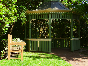 A green wooden gazebo with decorative trim is situated in a park or garden. In the foreground, there is a wooden bench placed on the grass. The gazebo is surrounded by lush green trees and plants, and sunlight creates dappled shadows on the ground.