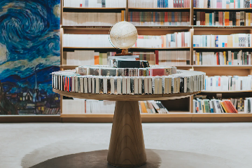 A modern library or bookstore interior featuring a round wooden table with books arranged standing up around the edge in a circle. On top of the table, there are stacked books and a globe. In the background, there are shelves filled with more books and a large mural resembling Van Gogh's Starry Night.