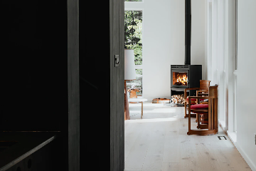 A modern, minimalist living space with light wooden floors, white walls, two wooden chairs with red cushions, a black wood-burning stove with a visible fire, a stack of firewood, and large windows allowing natural light to enter. Outside greenery is visible through the windows.