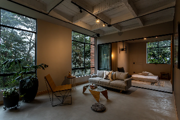 A modern living space with large windows showing a view of trees outside. The room features a light-colored sofa with decorative pillows, a geometric coffee table, and a contemporary chair. There are large potted plants near the windows, and a pouf on the floor. In the background, an adjoining bedroom with a low bed and minimalistic decor is visible. The room has industrial-style exposed ceilings and track lighting.