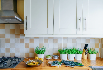 A modern kitchen countertop with several potted green plants, a cutting board with leafy greens, a frying pan containing cooked food, cups, a wooden knife block with knives and scissors, and a gas stove. The backsplash features beige and white tiles, and white cabinets are mounted above.