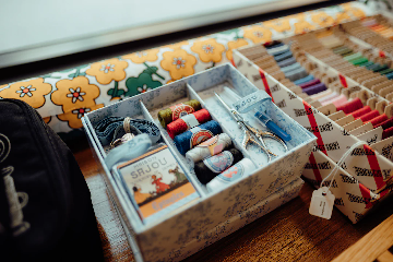 A close-up photo of a sewing kit on a wooden surface. The kit includes various colored threads, embroidery floss, small scissors shaped like a stork, a seam ripper, and a package of needles. There is also a box of assorted embroidery thread bobbins arranged by color in the background, and a piece of floral fabric with yellow, orange, and green flowers.
