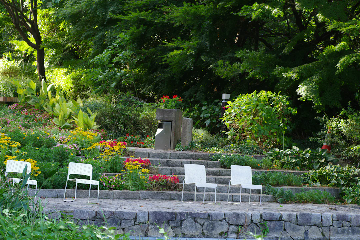 A garden scene with lush green plants, colorful flowers, and stone steps. Four white chairs are arranged on a stone patio in the foreground, facing away from the garden beds. There is a concrete trash bin and some red flowers on the steps in the background.