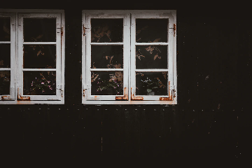 Two old, white-framed windows with visible rust and peeling paint on a dark wall. There are some potted plants with flowers inside the windows and floral-patterned curtains in the background.