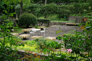 A peaceful outdoor garden area with greenery and flowering plants, featuring a round white metal table and three matching chairs. The table and chairs are surrounded by several square, low, yellow benches set on a paved section. Hedges and trimmed bushes border the space, and the atmosphere is serene and inviting.