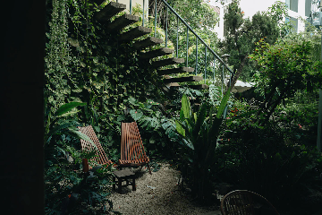 A lush, green outdoor space featuring two wooden lounge chairs and a small wooden table, surrounded by various tropical plants. There is an open wooden staircase with a metal railing leading upwards, and the area looks secluded and serene, ideal for relaxation.