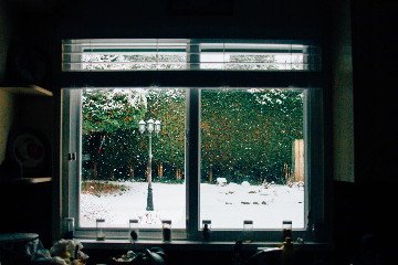 A view through a window from inside a house, showing a snowy backyard with falling snow, a street lamp, and tall green hedges in the background. The window sill has a few small bottles and kitchen items visible.