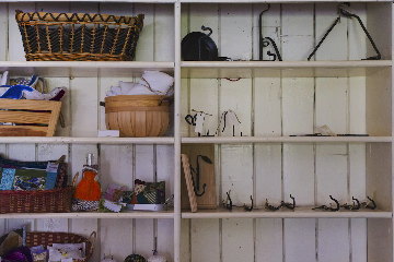 A set of wooden shelves attached to a white-paneled wall. The left shelves contain baskets, fabric, and handmade crafts including pillows, knitted items, and cloth pieces. The right shelves display metal hooks, a wooden bowl, and a black kettle, with many shelves partially empty except for iron hooks and hardware.